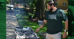 A man wearing a Mercy Home shirt prepares the grill for a barbecue.