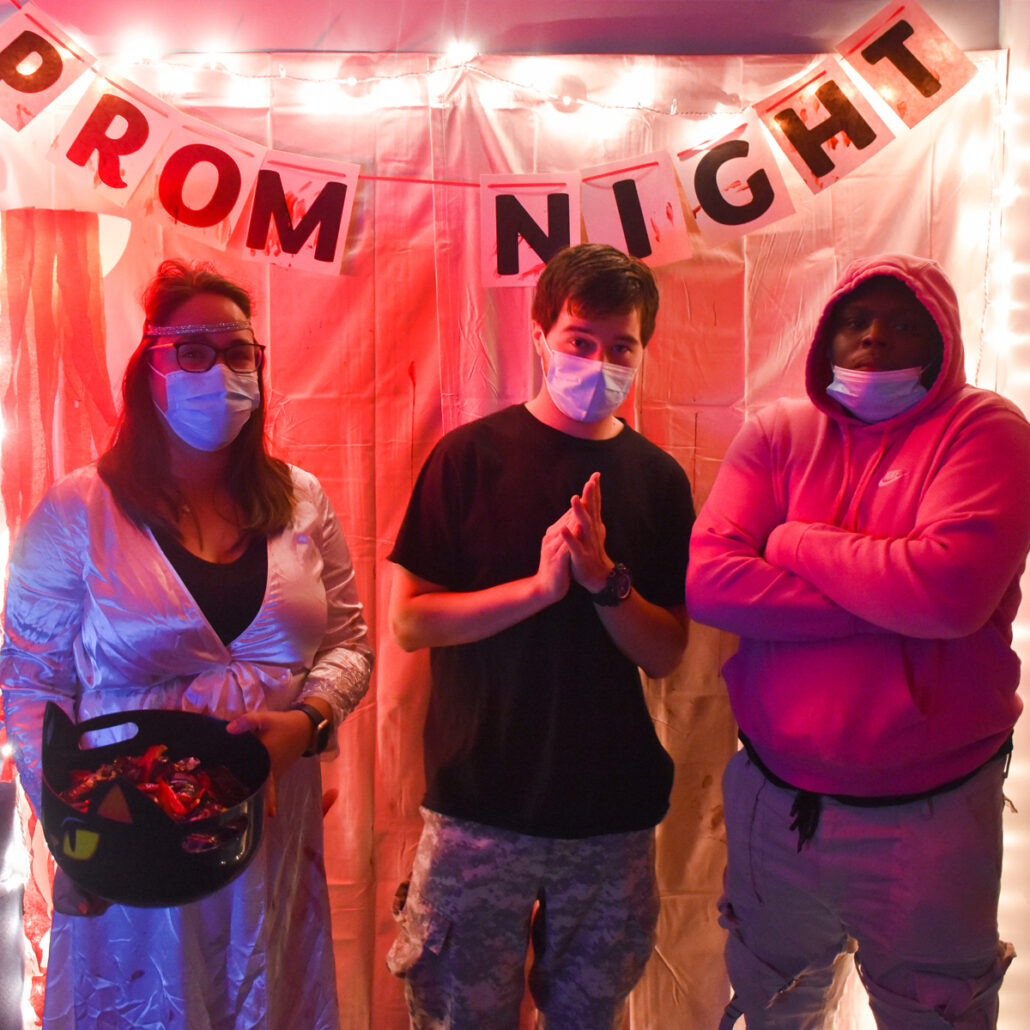 The words "Prom Night" hang from the ceiling in front of a white sheet illuminated in red. In front of the wall stands three people, enjoying the eerie school dance-themed room.