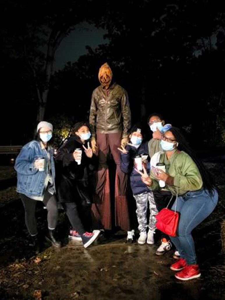 Mercy Home huddle together in front of a nighttime tree line for a group photo with a very tall pumpkin-headed monster from a haunted house. 
