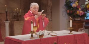 Very Rev. Gregory Sakowicz lifts his hands as he leads the Sunday Mass community in communion on Pentecost Sunday. He wears a red robe with yellow and orange patterning its front.
