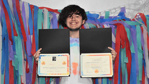 A young man holds up the two awards he received for his hard work during the school year.