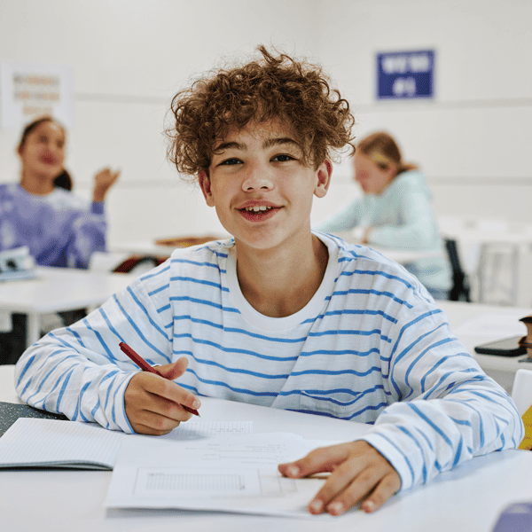 Teen boy in white and blue stripped shirt, sitting at his school desk with a pencil in his hand. Looking at viewer with a nice smile. 