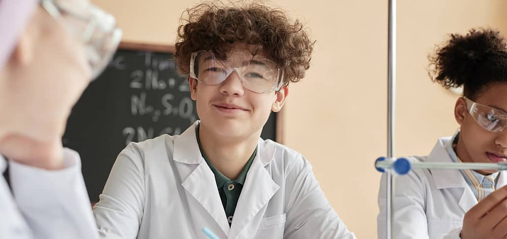 Close-up Vertical Portrait of a teenage boy, Alex, wearing a lab coat and protective lab glasses looking at the camera smiling.